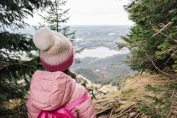 A girl in a pink jacket and beanie seen from behind looking out over the view of mountains and forests below on Alps Mountain