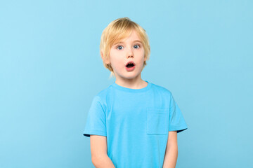 Wow! Portrait of a shocked cute little boy with blond hair covering open mouth with one hand, on pastel blue background. Surprised preschooler studio shot.