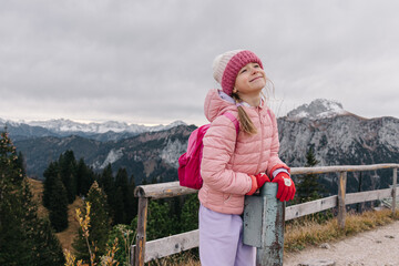 Happy young girl wearing pink jacket and beanie, standing on the wooden fence at mountain
