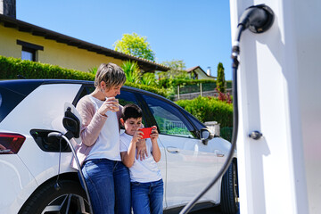 Son plays on phone while mom sips coffee, with their EV charging beside them.