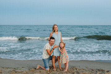 Naklejka premium Mother, father and child in love hugging on seashore. Happy family hugs a child barefoot standing on a sand beach. Man hugs pregnant wife. Dad, daughter kisses stomach mom enjoy summer vacation on sea