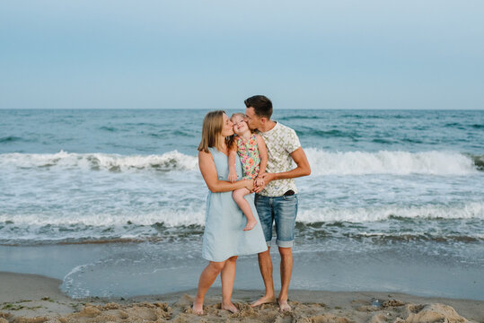 Mom, dad kisses daughter enjoy summer vacation on sea. Happy family hugs a child barefoot standing on water with big waves ocean near a sand beach. Mother, father and child in love hugging on seashore