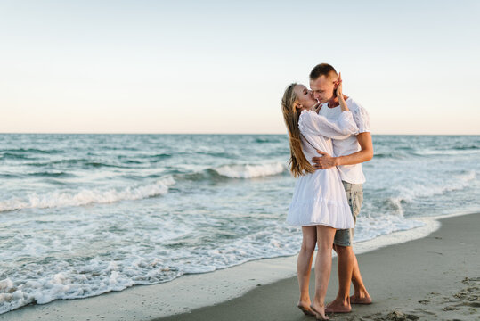 Couple in love hugging and kissing on seashore. Female hugs and kiss male stand barefoot on beach ocean and enjoy sunny summer day on vacation. Man embraces woman walk on sand sea. Spend time together
