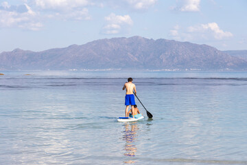 Naklejka premium A lone man enjoys paddleboarding on the tranquil blue waters with a mountainous backdrop, reflecting serenity and the simplicity of nature
