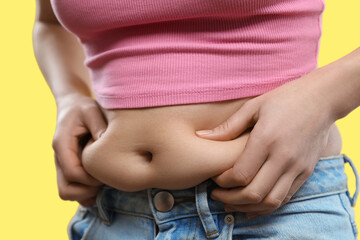 Woman touching belly fat on yellow background, closeup. Overweight problem - obrazy, fototapety, plakaty