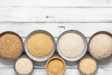Border of different varieties of uncooked rice in bowls on a white wooden background. View from above.