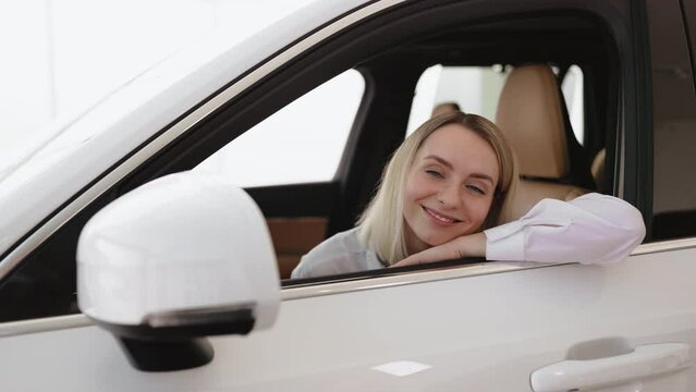 Calm woman, driver, customer, buyer, client in shirt putting hands on opened window, choose auto want buy new automobile in car showroom vehicle salon dealership store motor show indoor. Sales concept
