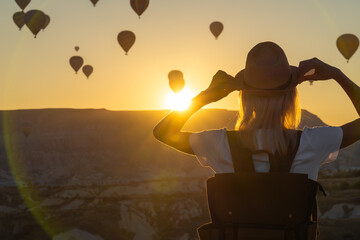 Young female tourist in a hat with a backpack admires the amazing scenery of hot air balloon flights over the valley of love at the moment of a beautiful sunrise, Cappadocia, Turkey.