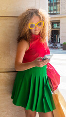 Coquettish s young female model in fashionable elegant bright multicolored green and red clothes looks at her phone against the background of the columns of an office building in the city. concept of