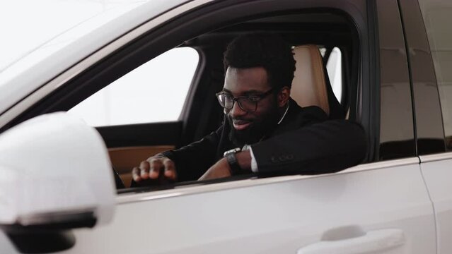 Calm African man, customer, buyer, client in suit holding hands on steering wheel choose auto want buy new automobile in car showroom vehicle salon dealership store motor show indoor. Sales concept.
