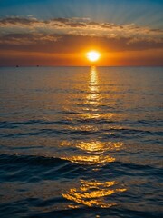 Fototapeta premium Serene sunset casts warm glow over calm sea. Suns reflection creates shimmering path on waters surface. Sky, adorned with hues of orange, yellow, blue.