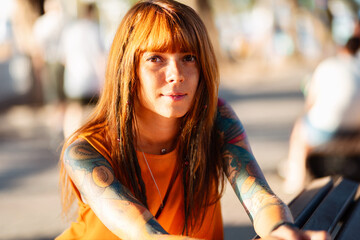 Portrait of young pretty Caucasian woman with tattoos poses sitting on bench in sunny light....