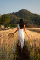 Back view of a woman in a white dress walking on a path through a wheat field, mountains in the background.