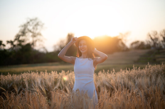 Joyful woman in a white dress holding a hat, standing in a wheat field at sunset, exuding happiness and freedom.
