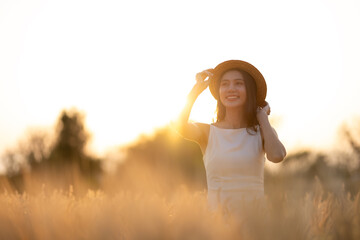 Obraz premium Joyful woman in a white dress holding a hat, standing in a wheat field at sunset, exuding happiness and freedom.