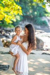 Young woman mother with a little daughter in white dresses on seashore in the shade of trees and palms