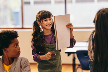 Happy young girl holding up a blank drawing book in a class