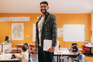 Happy elementary school teacher standing in a classroom, with children in the background
