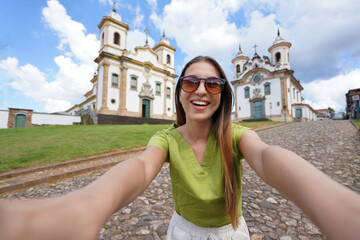 Holidays in Minas Gerais, Brazil. Beautiful traveler girl takes selfie picture with smartphone in Mariana, Minas Gerais, Brazil. - obrazy, fototapety, plakaty