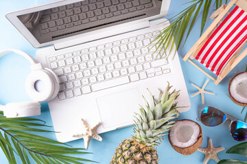 Work and vacation. Creative summer beach office flat lay with white laptop, tropical summer accessories, palm tree leaves, sunglasses, flip flops, pineapple and coconut on light blue background