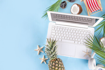 Work and vacation. Creative summer beach office flat lay with white laptop, tropical summer accessories, palm tree leaves, sunglasses, flip flops, pineapple and coconut on light blue background