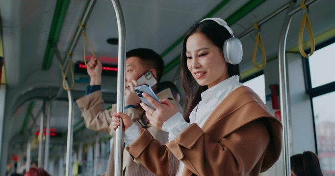 Cute Asian woman standing still next to handsome man. Wearing headphones and stylish coat. Listening to music while scrolling social media on smartphone. Browsing web. Searching for activities.