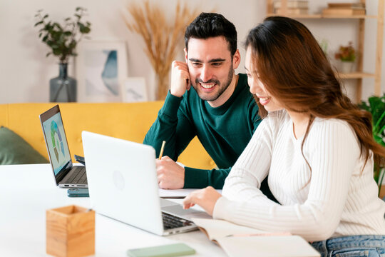 Two People Smiling And Working On Laptops At A Table.