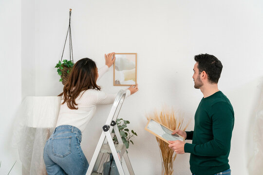 Woman hanging picture frame as man observes.