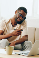 Young African American man working on his laptop while sitting on the sofa in his modern home He is a freelancer, smiling and typing with focus, surrounded by books and a cup of coffee The room is