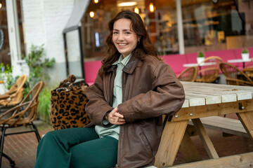 A smiling woman is sitting outside a cafe on a wooden bench, wearing a brown jacket over a green shirt and green trousers, holding her hands together.