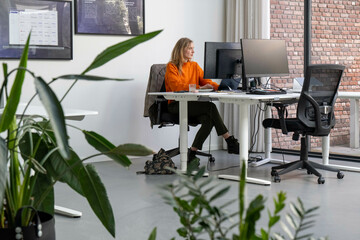 A woman is sitting at a desk in a modern office, working on a computer with dual monitors. She is wearing an orange top and black pants, and there is a plant nearby.