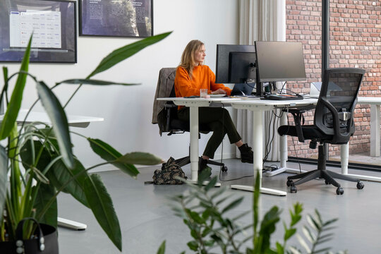 A woman is sitting at a desk in a modern office, working on a computer with dual monitors. She is wearing an orange top and black pants, and there is a plant nearby.