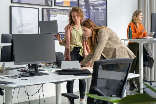 Two women are collaborating at a computer workstation in a modern office environment with another woman working at her desk in the background.