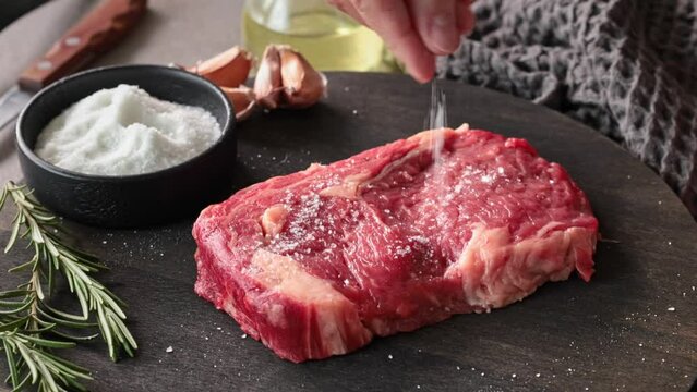 Chef sprinkles salt on fresh raw beef steak from both sides on kitchen table, close-up