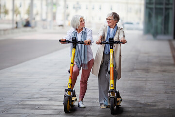 Mature joyful gray haired women friends ride electric scooters in the city, happy smiling, enjoying a fun time together