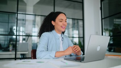 Online communication. Friendly brazilian or hispanic business woman, corporate manager, team leader, sits in the office, looks at laptop screen, makes greeting sign, brainstorming online with partners
