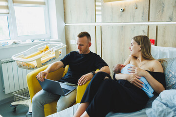 Young dad works online on a laptop while his wife plays with the baby. Young happy modern couple.