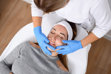 Dermatologist in white clothes and blue gloves makes a facial salon procedure for a girl with acne lying on a couch - 791695901