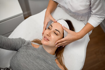 Facial massage for a woman in a sweater that lies on couch in a beauty salon - 791700924