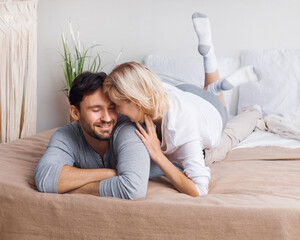 Portrait image - young playful couple looking at each other, lay on home bed. Blond woman, brunette...
