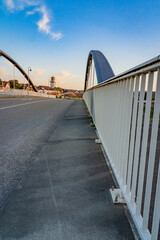 Die Brücke, eleganten Bögen und die glatte Oberfläche.  Straßenlaternen mit runden Köpfen beleuchtet die Brücke und folgt ihrer Krümmung. Das warme Leuchten des Sonnenuntergangs. Eine warme Atmosphäre