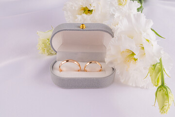 Two gold wedding classic rings in a gray velvet box on a white satin fabric with fresh white flowers. Wedding concept. Front view