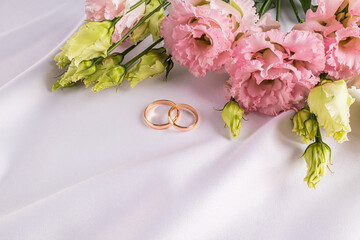 Two gold classic wedding rings on a white satin background with fresh pink austoma flowers. A copy space. postcard. invitation. cover.