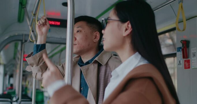 Cute Chinese couple standing next to each other. Holding by metal pole and safety strips for better balance. Woman pointing at something outside. Telling about city to her boyfriend.