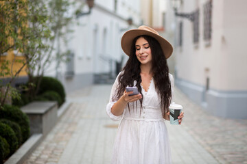 Naklejka premium Girl in a hat and white dress on the street. Young girl with coffee. Woman with long wavy hair in hat. Girl in a white dress and sneakers. Girl in the city takes a selfie.