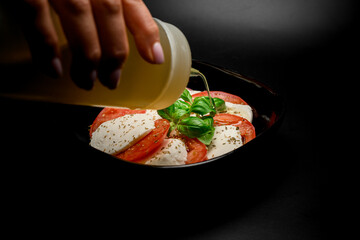 Woman's hand adds sauce in a bowl with tomatoes and soft cheese with greens - 791726724