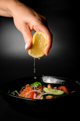 Female hand adds lemon juice to a black bowl with vegetable salad with bread croutons