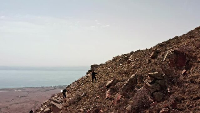 Climbers walking up a rocky slope to the top of a mountain, climbing over stones against the backdrop of a picturesque landscape and a lake in the distance.