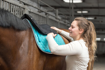 Young woman saddles the horse in the riding lane. She puts on the thick padded pad. The riding stable can be seen out of focus in the background.