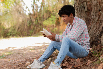 lifestyle in nature. young latin man sitting under a tree reading a book